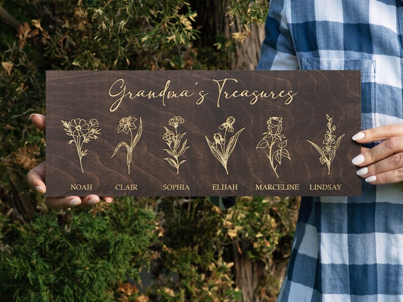 Puede incluir: Un letrero de madera marr&oacute;n oscuro con el texto "Grandma's Treasures" en letras doradas. Seis flores diferentes est&aacute;n grabadas en el letrero, cada una con un nombre debajo: Noah, Clair, Sophia, Elijah, Marceline y Lindsay.