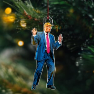 May include: A Christmas ornament featuring a cartoon illustration of a man in a blue suit and pink tie, with a red string for hanging.