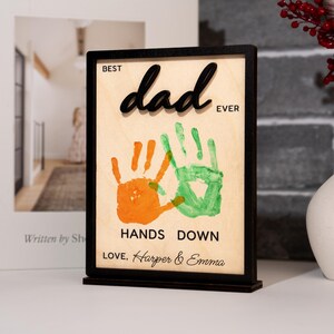 May include: A wooden sign with a black frame and base, featuring the words "Best Dad Ever" in a script font. Below are orange and green handprints, with the text "Hands Down" and the names "Love, Harper & Emma".