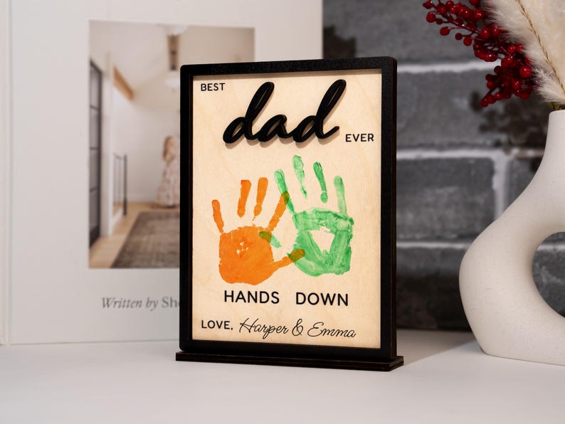 May include: A wooden sign with a black frame and base, featuring the words "Best Dad Ever" in a script font. Below are orange and green handprints, with the text "Hands Down" and the names "Love, Harper & Emma".