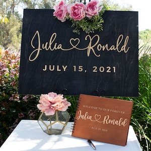 May include: A wooden sign with the names "Julia" and "Ronald" in gold script, a heart symbol, and the date "July 15, 2021". A wooden guest book with the same names and date is on a table with a pink flower in a glass vase.