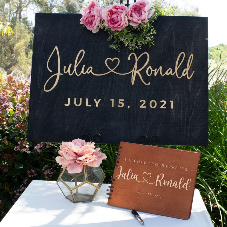 May include: A wooden sign with the names "Julia" and "Ronald" in gold script, a heart symbol, and the date "July 15, 2021". A wooden guest book with the same names and date is on a table with a pink flower in a glass vase.