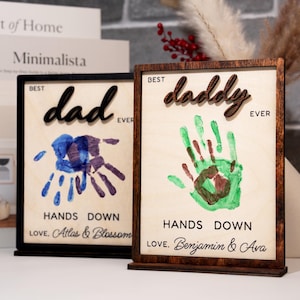 May include: Two wooden framed art pieces with the words "Best Dad Ever" and "Best Daddy Ever". Each features a handprint design in blue and green, respectively. The bottom of each frame reads "Hands Down" and a personalized message.