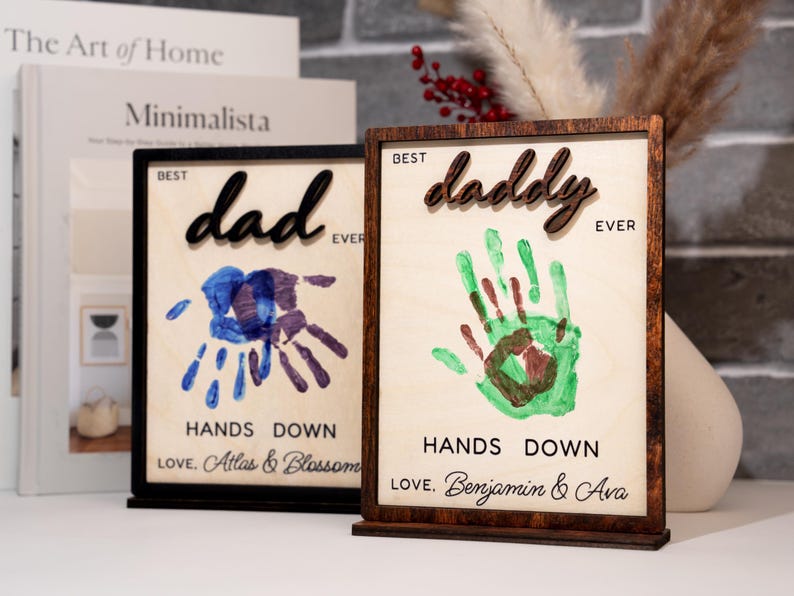 May include: Two wooden framed art pieces with the words "Best Dad Ever" and "Best Daddy Ever". Each features a handprint design in blue and green, respectively. The bottom of each frame reads "Hands Down" and a personalized message.