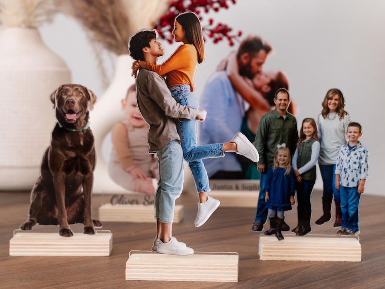 May include: Photo cutouts of a dog, a couple, a baby, and a family, each mounted on a wooden base. The couple is embracing, the family is smiling, and the dog is a chocolate lab. The baby is sitting.