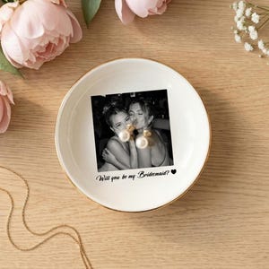 May include: A round white ceramic trinket dish with a gold-colored rim. The center features a black and white photograph of two people embracing, with the printed text "Will you be my Bridesmaid?" and a small black heart icon below the image.
