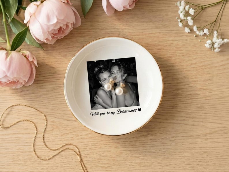 May include: A round white ceramic trinket dish with a gold-colored rim. The center features a black and white photograph of two people embracing, with the printed text "Will you be my Bridesmaid?" and a small black heart icon below the image.