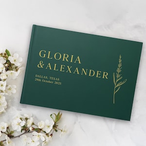 Puede incluir: Un libro de visitas de boda verde oscuro con letras doradas que dice "GLORIA & ALEXANDER" y la fecha "29 de octubre de 2025". Una ilustraci&oacute;n floral est&aacute; en el lado derecho. Flores blancas en el fondo.