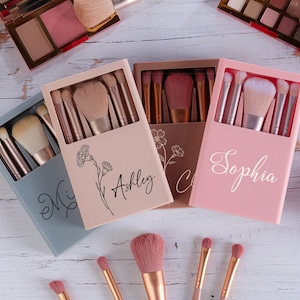 May include: Makeup brush sets in pastel-colored boxes, each containing multiple brushes. The boxes are personalized with names in script. The image also shows eyeshadow palettes and individual makeup brushes.
