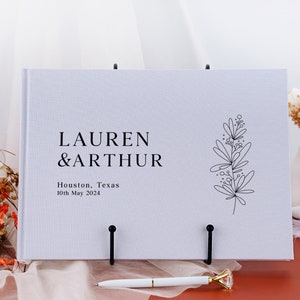 Puede incluir: Un libro de visitas de boda blanco con los nombres "LAUREN & ARTHUR" impresos en negro. El libro tambi&eacute;n incluye la ubicaci&oacute;n "Houston, Texas" y la fecha "10 de mayo de 2024". Una ilustraci&oacute;n floral negra est&aacute; en el lado derecho.