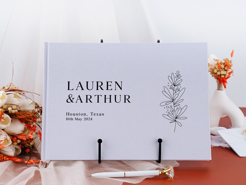 Puede incluir: Un libro de visitas de boda blanco con los nombres "LAUREN & ARTHUR" impresos en negro. El libro tambi&eacute;n incluye la ubicaci&oacute;n "Houston, Texas" y la fecha "10 de mayo de 2024". Una ilustraci&oacute;n floral negra est&aacute; en el lado derecho.