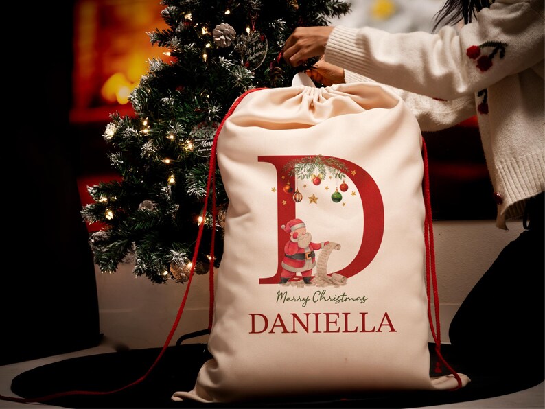 May include: A white Christmas gift bag with a red letter D and a Santa Claus illustration. The bag has a red drawstring and the text "Merry Christmas Daniella" is printed on the front.