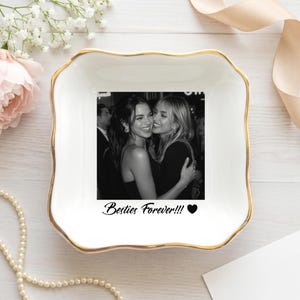 May include: A square white ceramic trinket dish with scalloped edges and a gold-painted rim. The center features a black and white photograph of two people embracing, with the text "Besties Forever!!!" and a black heart icon printed below.
