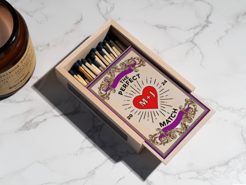 Puede incluir: Una caja de madera de cerillas con una etiqueta personalizada que dice "The Perfect Match" con un coraz&oacute;n que dice "M+J" y los n&uacute;meros "20" y "24" a cada lado del coraz&oacute;n. La etiqueta tiene un dise&ntilde;o dorado y morado.