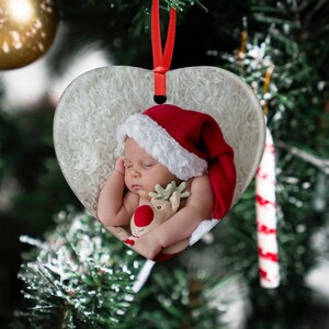 May include: A white heart-shaped Christmas ornament with a sleeping baby wearing a red Santa hat and holding a plush reindeer toy.