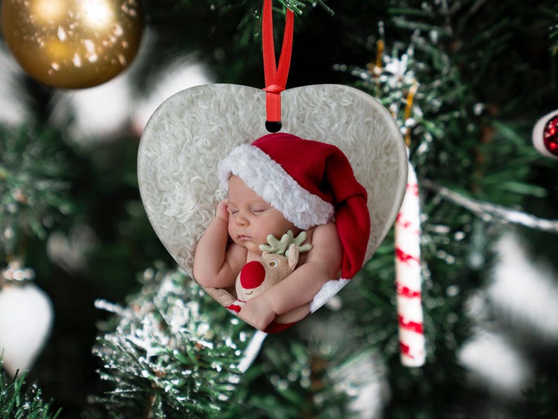 May include: A white heart-shaped Christmas ornament with a sleeping baby wearing a red Santa hat and holding a plush reindeer toy.