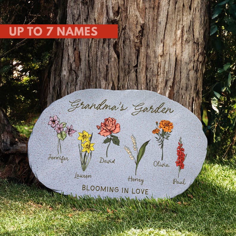 May include: A gray stone garden marker with the text "Grandma's Garden" and the phrase "Blooming in Love." It features colorful illustrations of flowers and names: Jennifer, Lauren, David, Henry, Olivia, and Paul. The marker can accommodate up to 7 names.