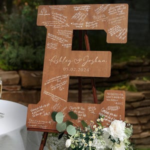 K&ouml;nnte beinhalten: Ein gro&szlig;es Holzschild in Form des Buchstabens "E" mit den Namen "Ashley & Joshua" und dem Datum "05.02.2024" eingraviert. Das Schild ist mit handgeschriebenen Nachrichten bedeckt und mit Blumen und Gr&uuml;n geschm&uuml;ckt. Es wird auf einer Staffelei ausgestellt.