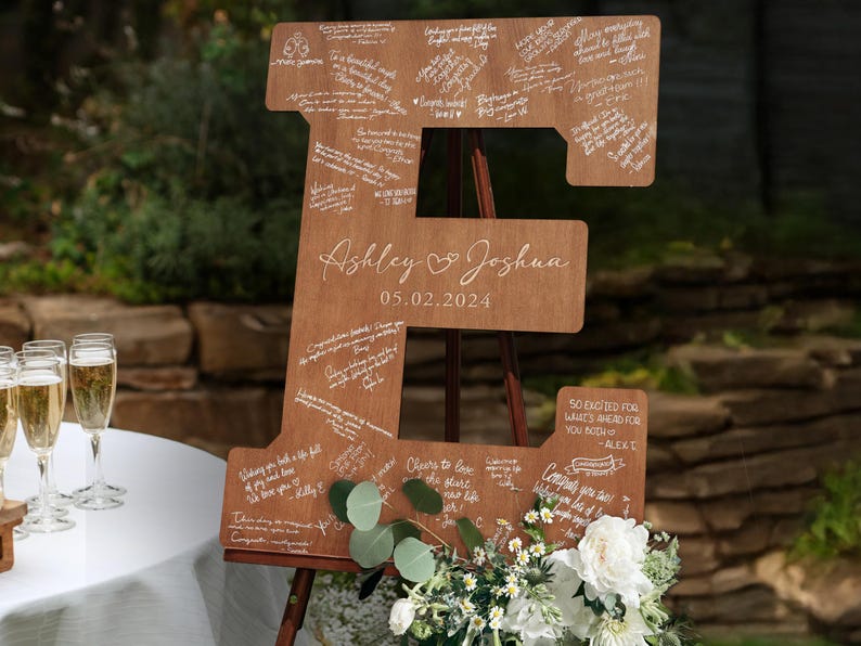 K&ouml;nnte beinhalten: Ein gro&szlig;es Holzschild in Form des Buchstabens "E" mit den Namen "Ashley & Joshua" und dem Datum "05.02.2024" eingraviert. Das Schild ist mit handgeschriebenen Nachrichten bedeckt und mit Blumen und Gr&uuml;n geschm&uuml;ckt. Es wird auf einer Staffelei ausgestellt.