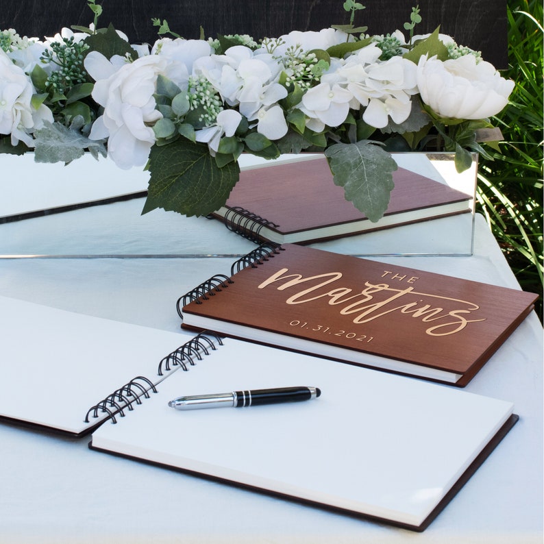 May include: Two brown wooden guest books with white pages and gold lettering. The top book says "The Martins 01.31.2021". A black pen is lying on the open pages of the bottom book.