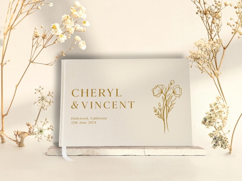 Puede incluir: Un libro de visitas de boda color crema con letras doradas que dice "CHERYL & VINCENT". El libro incluye la fecha y el lugar de la boda. Un dise&ntilde;o floral dorado est&aacute; en el lado derecho. Una cinta blanca de marcador es visible.