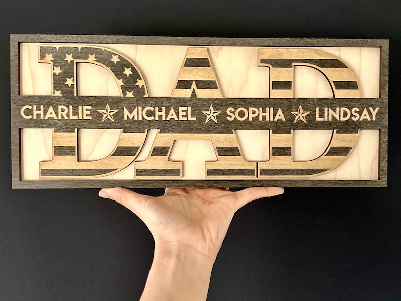 May include: Wooden sign with the word "DAD" in large letters, each letter is made of wood with an American flag design. The sign has a dark brown frame and a light wood background. The names "CHARLIE", "MICHAEL", "SOPHIA", and "LINDSAY" are engraved below the letters.