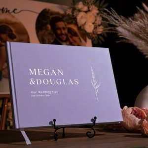 Puede incluir: Un libro de visitas de boda lila con los nombres "Megan & Douglas" y la fecha "13 de octubre de 2024" impresos en blanco. El libro se exhibe sobre un soporte de metal negro, con una ilustraci&oacute;n floral. Un jarr&oacute;n con hierba de la pampa y flores est&aacute; en el fondo.