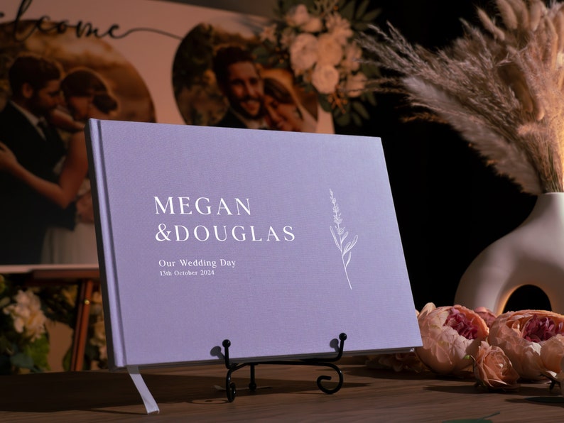 Puede incluir: Un libro de visitas de boda lila con los nombres "Megan & Douglas" y la fecha "13 de octubre de 2024" impresos en blanco. El libro se exhibe sobre un soporte de metal negro, con una ilustraci&oacute;n floral. Un jarr&oacute;n con hierba de la pampa y flores est&aacute; en el fondo.