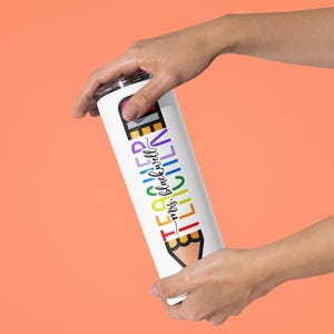 May include: White insulated tumbler with a colorful pencil design and the text "TEACHER mrs. blackwell". The tumbler is held against an orange background. The design features rainbow colors.