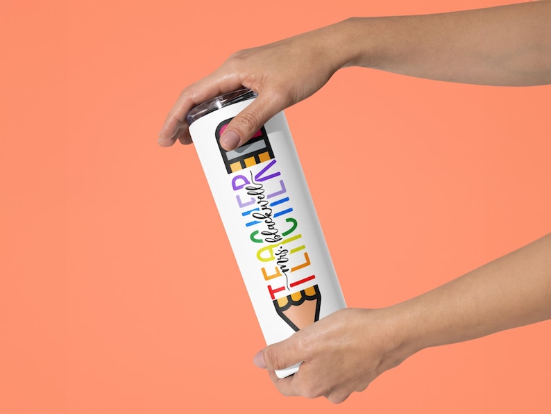 May include: White insulated tumbler with a colorful pencil design and the text "TEACHER mrs. blackwell". The tumbler is held against an orange background. The design features rainbow colors.
