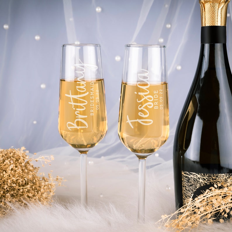 Puede incluir: Dos copas de champ&aacute;n llenas de un l&iacute;quido dorado, personalizadas con nombres y fechas. Una dice "Brittany, Bridesmaid 05.09.22" y la otra "Jessica, Bride 05.09.22". Una botella de champ&aacute;n es parcialmente visible.