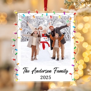 Pode incluir: Enfeite de Natal em acr&iacute;lico transparente com uma foto de fam&iacute;lia no interior, emoldurado por uma sequ&ecirc;ncia de luzes coloridas. A foto mostra uma fam&iacute;lia de quatro pessoas com um boneco de neve em um cen&aacute;rio de neve. O texto diz "The Anderson Family 2025."