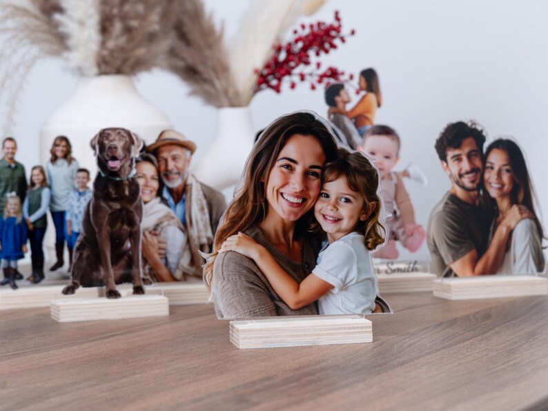 May include: Family photo cutouts on wooden bases, featuring various family members and pets. The cutouts are displayed on a wooden surface, with a neutral background and decorative vases.