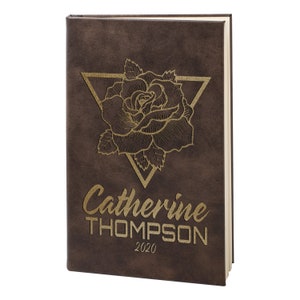 May include: A brown leather-bound journal with a gold embossed rose design and a triangle. The journal is personalized with the name "Catherine THOMPSON" and the year "2020".