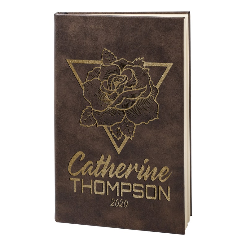 May include: A brown leather-bound journal with a gold embossed rose design and a triangle. The journal is personalized with the name "Catherine THOMPSON" and the year "2020".
