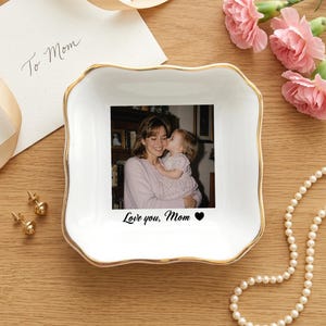 May include: A square white ceramic trinket dish with a gold-trimmed scalloped edge. The center features a black and white photograph of a person holding a child, with the printed text Love you, Mom and a black heart icon below the image.