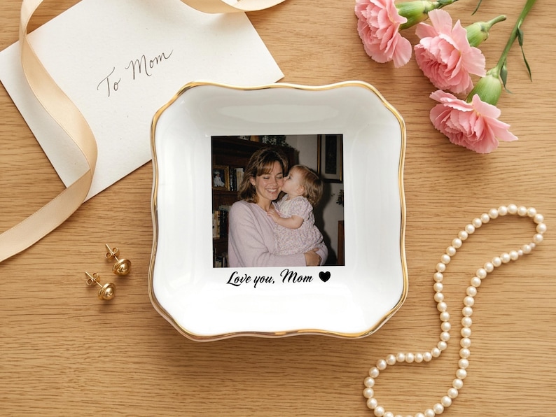 May include: A square white ceramic trinket dish with a gold-trimmed scalloped edge. The center features a black and white photograph of a person holding a child, with the printed text Love you, Mom and a black heart icon below the image.