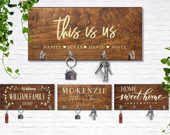 Può includere: Quattro portachiavi da parete rettangolari in legno scuro con ganci argentati. I cartelli presentano scritte bianche e dorate tra cui "this is us", "WILLIAM FAMILY", "MC KENZIE" e "HOME sweet home", insieme a vari nomi e date.