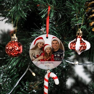May include: A white ceramic Christmas ornament with a photo of a family of three. The ornament is round and has a red ribbon attached to the top. The photo shows a woman, a man, and a young girl wearing Santa hats. The text "Stewart Family 2024" is printed on the ornament.