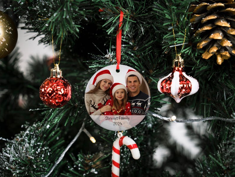May include: A white ceramic Christmas ornament with a photo of a family of three. The ornament is round and has a red ribbon attached to the top. The photo shows a woman, a man, and a young girl wearing Santa hats. The text "Stewart Family 2024" is printed on the ornament.
