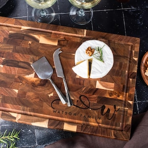 May include: A wooden chopping board with a dark brown, end-grain pattern. It features a carved juice groove and the text "WATSON FAMILY" with a stylized "W". A cheese knife and spatula rest on the board, alongside brie cheese and crackers.