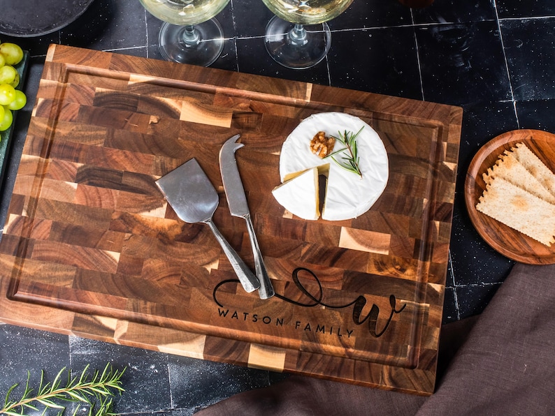 May include: A wooden chopping board with a dark brown, end-grain pattern. It features a carved juice groove and the text "WATSON FAMILY" with a stylized "W". A cheese knife and spatula rest on the board, alongside brie cheese and crackers.