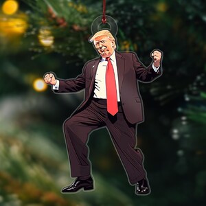 May include: A Christmas ornament featuring a cartoon illustration of a man in a dark suit and red tie, with a white shirt and blonde hair, striking a celebratory pose with his arms raised.