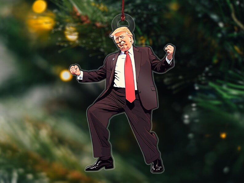 May include: A Christmas ornament featuring a cartoon illustration of a man in a dark suit and red tie, with a white shirt and blonde hair, striking a celebratory pose with his arms raised.