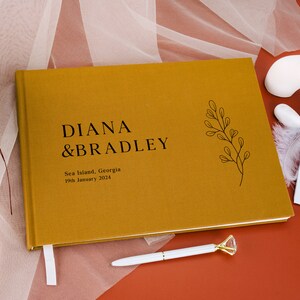 Puede incluir: Libro de visitas de boda de color mostaza con los nombres "DIANA & BRADLEY" en texto negro. El libro tambi&eacute;n incluye la ubicaci&oacute;n "Sea Island, Georgia" y la fecha "19 de enero de 2024". Tambi&eacute;n son visibles una cinta de marcador blanca y un bol&iacute;grafo con una parte superior en forma de diamante.