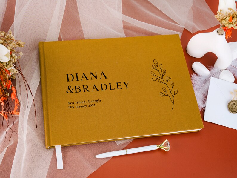 Puede incluir: Libro de visitas de boda de color mostaza con los nombres "DIANA & BRADLEY" en texto negro. El libro tambi&eacute;n incluye la ubicaci&oacute;n "Sea Island, Georgia" y la fecha "19 de enero de 2024". Tambi&eacute;n son visibles una cinta de marcador blanca y un bol&iacute;grafo con una parte superior en forma de diamante.