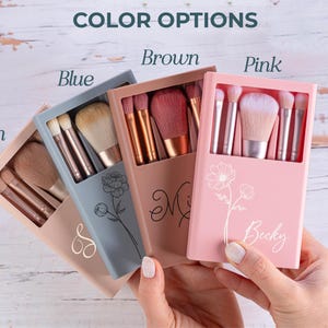 May include: A collection of makeup brush sets in tan, blue, brown, and pink cases. Each case is personalized with a name or initial and features a floral design. The cases are open, revealing the brushes inside.