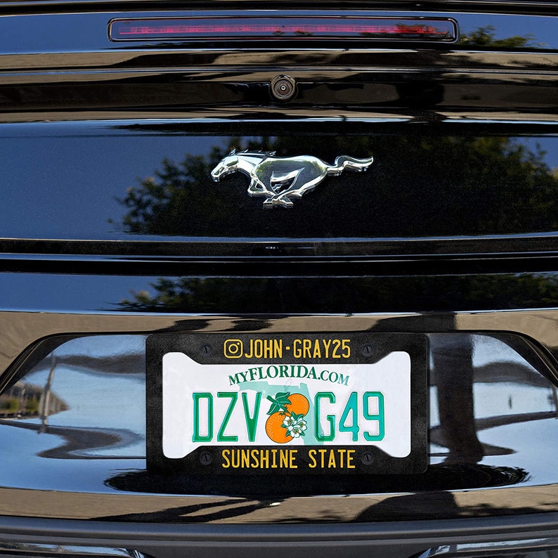 Puede incluir: Un coche negro con un logotipo de Mustang plateado en la parte trasera. El coche tiene una matr&iacute;cula de Florida con el texto "DZV G49" y las palabras "MYFLORIDA.COM" y "SUNSHINE STATE" en ella. El marco de la matr&iacute;cula tiene el texto "@JOHN-GRAY25" en &eacute;l.