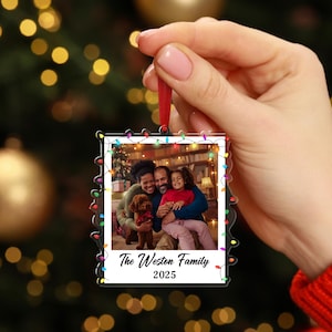 Pode incluir: Enfeite de Natal em acr&iacute;lico transparente com uma foto de fam&iacute;lia no interior. O enfeite tem a forma de uma foto polaroid e &eacute; adornado com luzes coloridas. O texto "The Weston Family 2025" est&aacute; impresso na parte inferior.