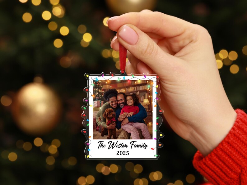 Pode incluir: Enfeite de Natal em acr&iacute;lico transparente com uma foto de fam&iacute;lia no interior. O enfeite tem a forma de uma foto polaroid e &eacute; adornado com luzes coloridas. O texto "The Weston Family 2025" est&aacute; impresso na parte inferior.